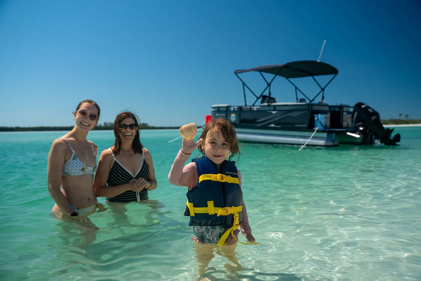 A family standing in knee-deep clear water beside the anchored pontoon boat